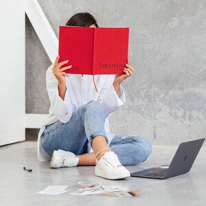 May include: A person is sitting on the floor, holding a red notebook with the word "STRATEGY" visible. They are wearing a white shirt, blue jeans, and white sneakers. A laptop and papers are nearby.