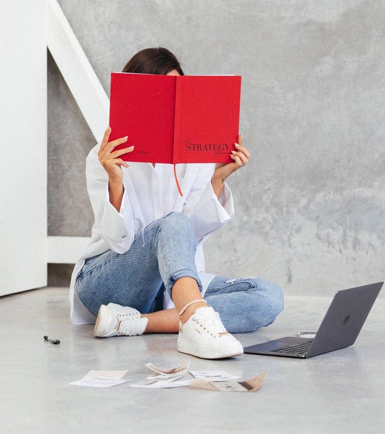 May include: A person is sitting on the floor, holding a red notebook with the word "STRATEGY" visible. They are wearing a white shirt, blue jeans, and white sneakers. A laptop and papers are nearby.