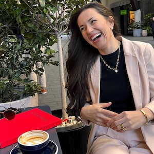 May include: A woman wearing a black top and a light pink blazer laughs while sitting at an outdoor table. On the table are a red book, sunglasses, a smartphone, and a cup of coffee. A gold necklace and bracelet are visible.