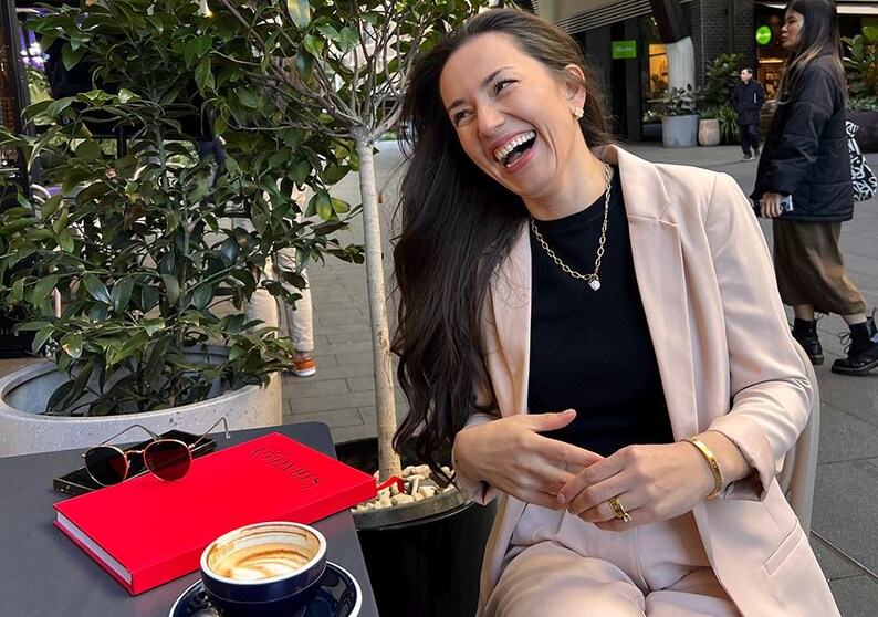 May include: A woman wearing a black top and a light pink blazer laughs while sitting at an outdoor table. On the table are a red book, sunglasses, a smartphone, and a cup of coffee. A gold necklace and bracelet are visible.