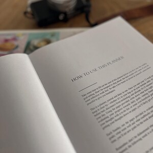 May include: Open planner with the text "HOW TO USE THIS PLANNER" in a serif font. The pages are white with black text. A camera is out of focus in the background. The planner is open on a wooden surface.