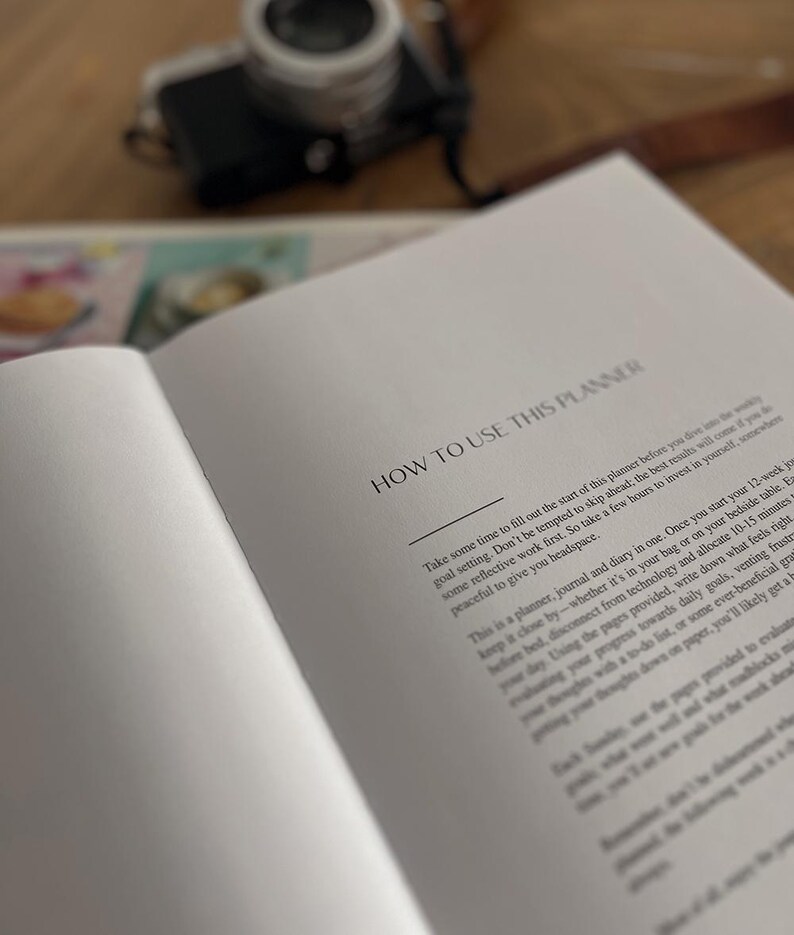 May include: Open planner with the text "HOW TO USE THIS PLANNER" in a serif font. The pages are white with black text. A camera is out of focus in the background. The planner is open on a wooden surface.