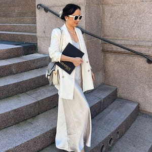 May include: A woman wearing a white blazer, sunglasses, and a cream-colored dress, holding a black notebook with the word "STRATEGY" printed on it. She is standing on a set of stone steps, with a purse over her shoulder.