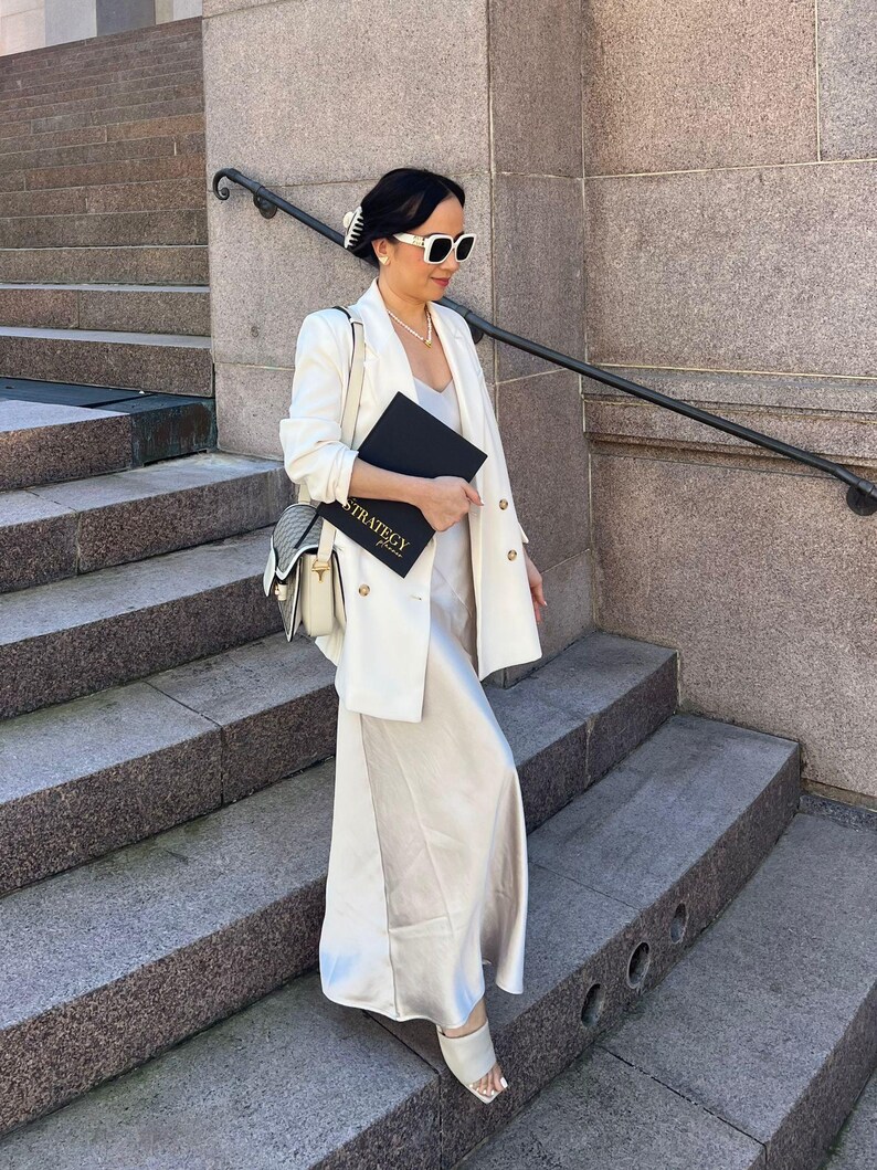 May include: A woman wearing a white blazer, sunglasses, and a cream-colored dress, holding a black notebook with the word "STRATEGY" printed on it. She is standing on a set of stone steps, with a purse over her shoulder.