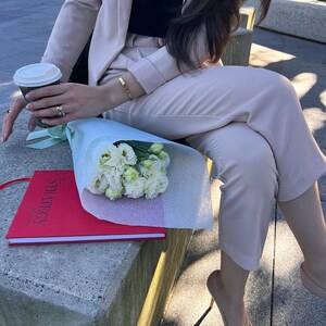 May include: A light pink pantsuit with a black top, paired with nude heels. A red notebook with the word "STRATEGY" is visible, along with a bouquet of white flowers and a coffee cup. A gold bracelet and rings complete the look.