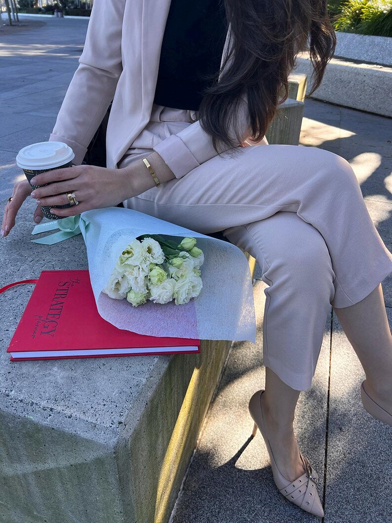 May include: A light pink pantsuit with a black top, paired with nude heels. A red notebook with the word "STRATEGY" is visible, along with a bouquet of white flowers and a coffee cup. A gold bracelet and rings complete the look.