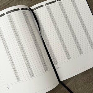 May include: Open black planner with a ribbon bookmark, displaying a weekly schedule layout. Each day is divided into time slots from 6:00 AM to 9:00 PM. The planner is open on a wooden surface.