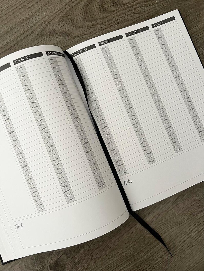 May include: Open black planner with a ribbon bookmark, displaying a weekly schedule layout. Each day is divided into time slots from 6:00 AM to 9:00 PM. The planner is open on a wooden surface.