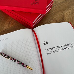 May include: Red hardcover planner with the text "the STRATEGIC planner" stacked on top of each other. An open planner with a quote and a floral pen are on the right. The quote reads, "I NEVER DREAMED ABOUT SUCCESS. I WORKED FOR".