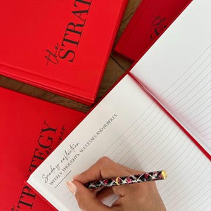 May include: Red "The Strategy Planner" notebooks with black script text. One notebook is open, revealing lined pages. A pen with a black, pink, and gold design is writing on the open page. The text "Sunday reflection" and "Weekly thoughts, successes and hurdles" is visible.