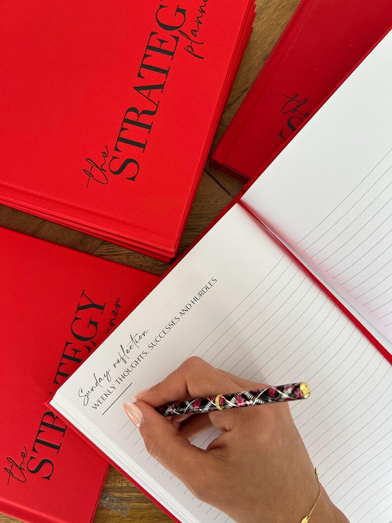 May include: Red "The Strategy Planner" notebooks with black script text. One notebook is open, revealing lined pages. A pen with a black, pink, and gold design is writing on the open page. The text "Sunday reflection" and "Weekly thoughts, successes and hurdles" is visible.