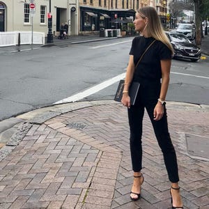 May include: A person wearing a black t-shirt, black pants, and black high-heeled sandals stands on a brick-paved street corner. They are holding a black book. The background includes buildings, cars, and a crosswalk.