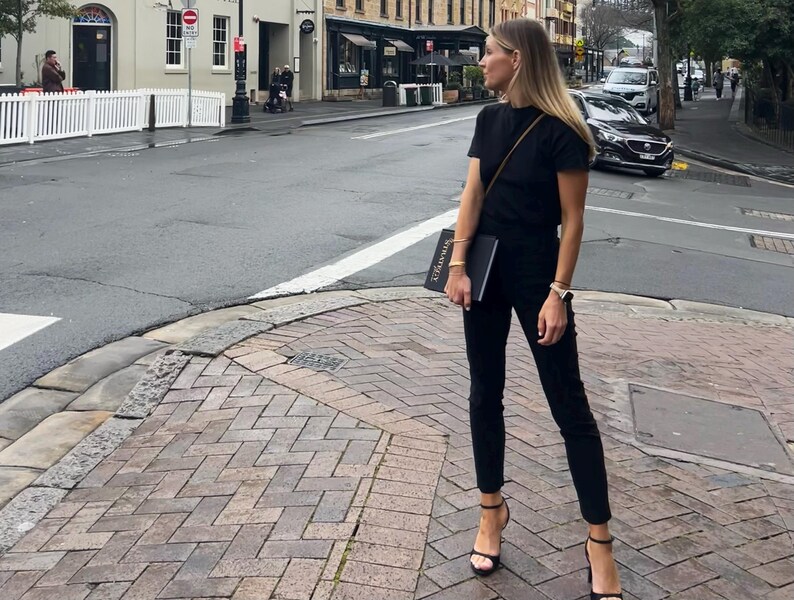 May include: A person wearing a black t-shirt, black pants, and black high-heeled sandals stands on a brick-paved street corner. They are holding a black book. The background includes buildings, cars, and a crosswalk.