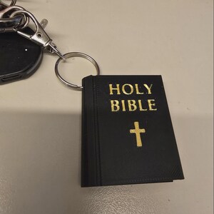 May include: A black Holy Bible keyring with gold lettering and a gold cross. The keyring is attached to a silver key ring and a car key fob.