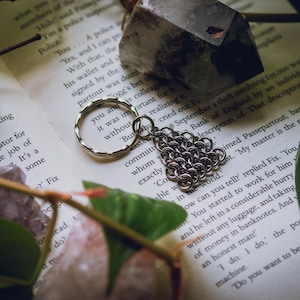 May include: A silver-toned metal keyring featuring a circular ring and a triangular charm constructed from interlocking chainmail rings. The keyring is positioned on an open book page with visible printed text, surrounded by natural elements.