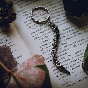 May include: A silver-toned metal keyring featuring a circular ring attached to a long, flexible chain made of interlocking jump rings, ending in a sharp, pointed metal spike. The chain has a textured, woven appearance with a metallic finish.