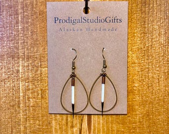 May include: Handmade Alaskan earrings featuring teardrop-shaped brass hoops with a white and black quill accent. The earrings are displayed on a brown tag with the text "ProdigalStudioGifts Alaskan Handmade". The background is a wooden surface.