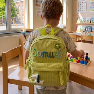 May include: A light green corduroy backpack with the name "Samuel" embroidered in blue and green thread. The backpack features a flower applique and a dog patch. It has a top handle and adjustable straps.