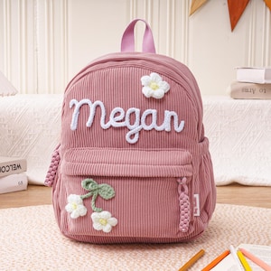 May include: A pink corduroy backpack with the name "Megan" embroidered in white. The backpack features white flower and green leaf accents, a small front pocket, and a top handle. A small alarm clock and books are in the background.