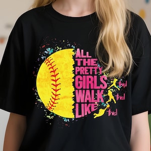 May include: Black t-shirt featuring a softball design with the text "All the Pretty Girls Walk Like This!". The design includes a yellow softball, pink text, and colorful splatter accents.