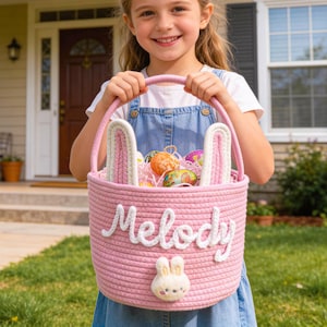 以下が含まれることがあります： ピンク色のイースターバスケット。ウサギの耳と白い筆記体で「Melody」と書かれています。バスケットにはカラフルなイースターエッグと装飾用の紙が入っています。小さなウサギの顔が前面に付いています。