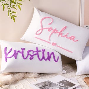 Könnte beinhalten: Zwei weiße, rechteckige Kissen mit gestickten Namen. Ein Kissen hat den Namen "Sophia" in rosa Schrift mit einem Herz. Das andere Kissen hat den Namen "Kristin" in lila Schrift. Die Kissen liegen auf einem Bett.