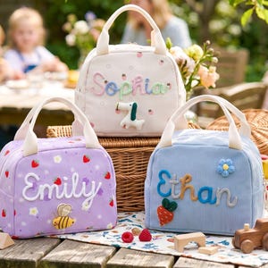 May include: Three corduroy lunch bags with top handles and zipper closures. The bags are cream, lavender, and light blue, each featuring embroidered names: Sophia, Emily, and Ethan. Decorative patches include a dog, a bee, a flower, and a carrot.