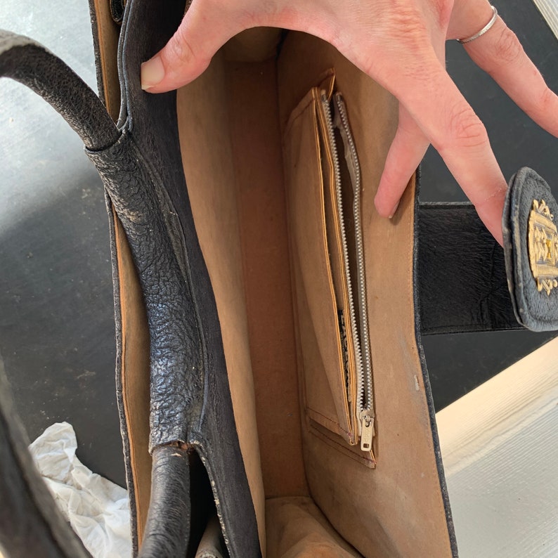 May include: Black leather handbag with a zippered pocket and a gold metal accent. The bag has a brown lining and a top handle.