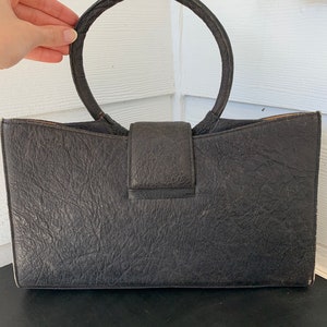 May include: A black leather handbag with a large, circular handle. The bag has a textured surface and a flap closure.