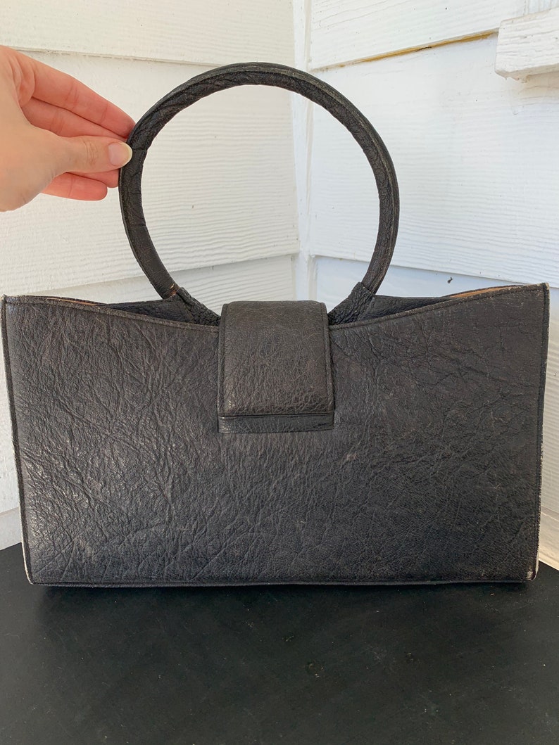 May include: A black leather handbag with a large, circular handle. The bag has a textured surface and a flap closure.