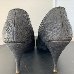 May include: A pair of black leather heeled shoes with a textured finish. The heels are thin and have a gold accent at the bottom.