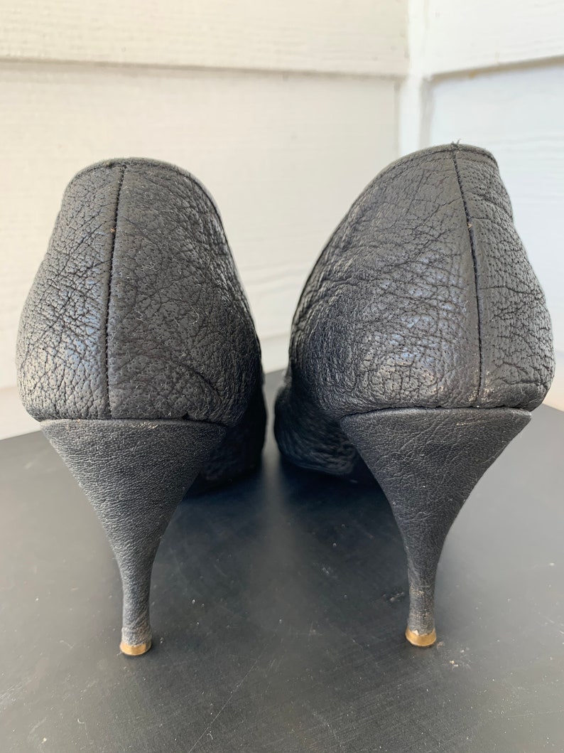 May include: A pair of black leather heeled shoes with a textured finish. The heels are thin and have a gold accent at the bottom.