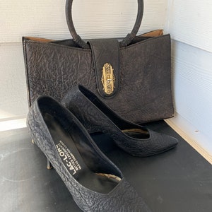 May include: A black leather handbag with a gold-toned clasp and a circular handle. The handbag is sitting on a wooden surface next to a pair of black leather shoes with a pointed toe.