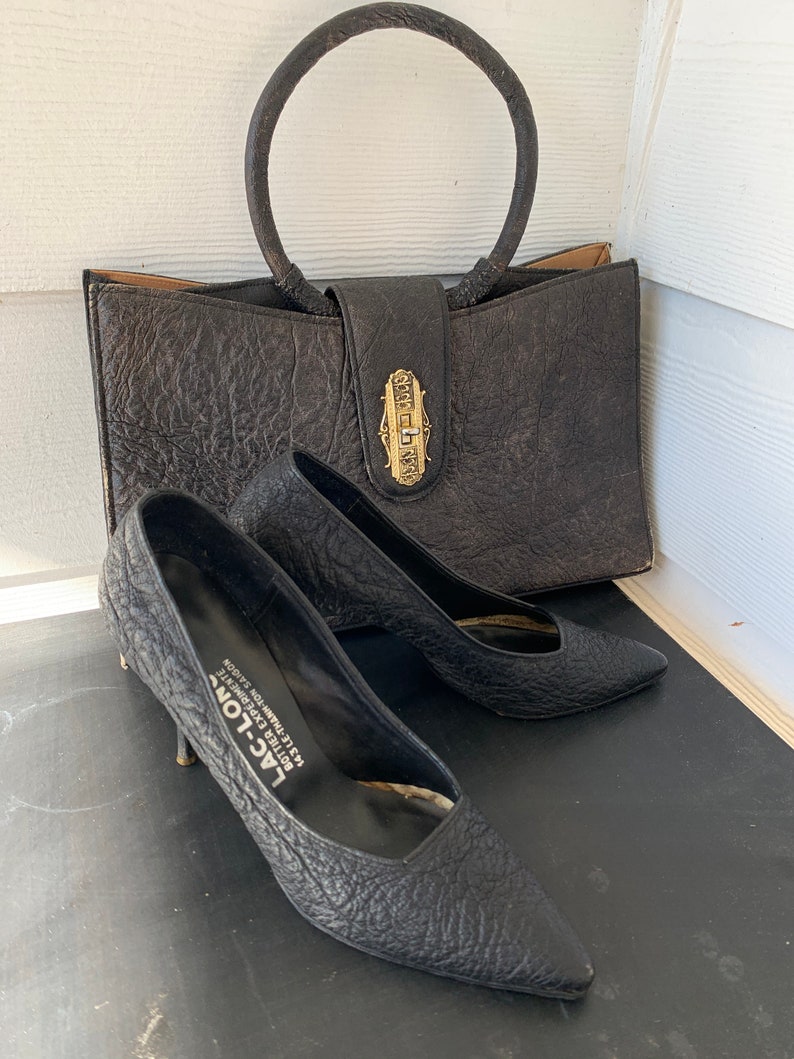 May include: A black leather handbag with a gold-toned clasp and a circular handle. The handbag is sitting on a wooden surface next to a pair of black leather shoes with a pointed toe.