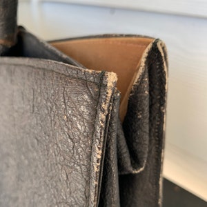 May include: Close-up of a black leather tote bag with a brown leather lining. The bag has a worn, distressed look, with visible stitching and creases.