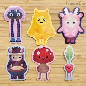 May include: A set of six holographic stickers featuring various characters: a tall dark creature, a yellow grumpy figure, a pink skull-themed being, a dark brown horned creature, a red mushroom character, and a small pink radish with green leaves.