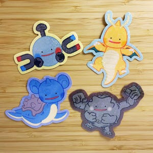May include: Four colorful stickers featuring cartoon characters. One is a blue and silver magnet with a red mouth. Another is a yellow dragon-like creature. A blue sea creature and a gray rock-like character complete the set.