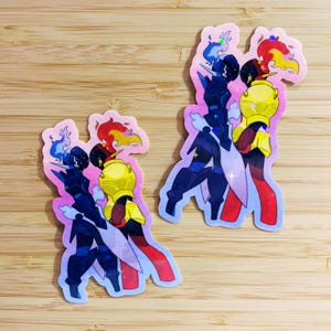 May include: Two stickers featuring stylized figures with flame and energy effects. One figure is primarily blue with white accents, the other is yellow and red. The stickers have a pink outline and are on a wooden surface.