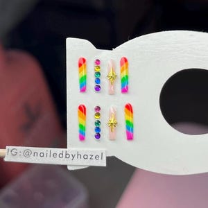 Pode incluir: Conjunto de unhas postiças com um design de arco-íris, pedras preciosas coloridas e detalhes de estrelas douradas. As unhas são exibidas em um cartão branco. O texto no cartão diz "G: @nailedbyhazel".