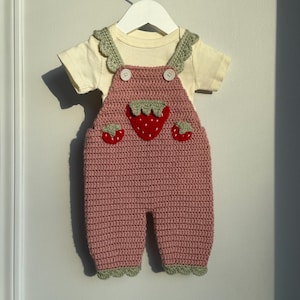 May include: A handmade baby outfit, including a light yellow short-sleeved shirt and a pink crocheted overall. The overall features a large strawberry applique and two smaller strawberry accents. The edges are trimmed with green crochet.