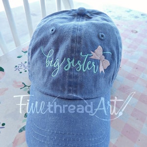 May include: A light blue denim baseball cap with the text "big sister" embroidered in green and a pink bow.