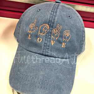 May include: A blue baseball cap with the word "LOVE" spelled out in American Sign Language, embroidered in orange thread. The cap has a curved brim and a fabric strap for adjustment.