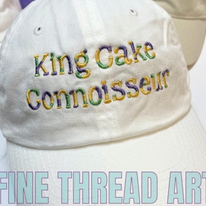 May include: White baseball cap with embroidered text "King Cake Connoisseur" in purple, green, and gold thread. The text "FINE THREAD ART" is printed in purple and blue on the brim of the cap.