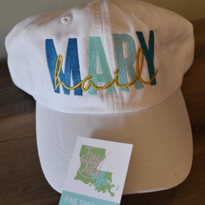 May include: White baseball cap with the name "MARY" embroidered in blue, teal, and gold thread. The cap has a curved brim and a small logo on the back that says "Fine Thread Art Embroidery".