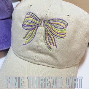 May include: A beige baseball cap with an embroidered purple, green, and yellow bow. The text "FINE THREAD ART" is on the front of the cap.