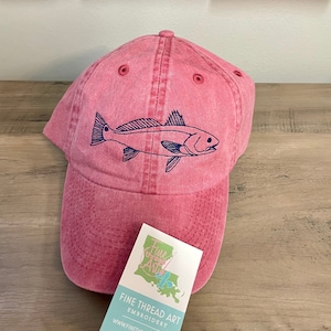 May include: A pink baseball cap with a blue embroidered redfish design. The cap has a distressed look and is made of cotton. The cap is sitting on a wooden surface with a business card that says "Fine Thread Art Embroidery" and a website address.