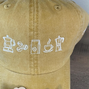 May include: A mustard yellow baseball cap with white embroidered coffee-themed designs. The cap has a curved brim and a white embroidered design of a coffee pot, coffee beans, a bag of coffee, a cup of coffee, and a Moka pot. The cap is sitting on a wooden surface with a wooden sign that says "fine thread art embroidery".