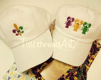 May include: Two white baseball caps with embroidered designs. One cap features a fleur-de-lis in green, gold, and purple. The other cap has three embroidered lobsters in red, yellow, and green.