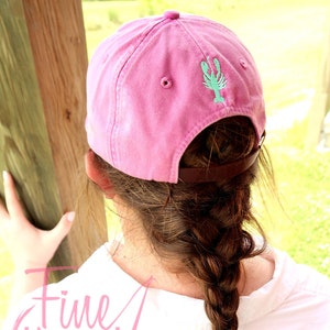 May include: Pink baseball cap with a green and blue embroidered lobster design. The cap has a brown leather strap and is worn by a person with long brown hair.