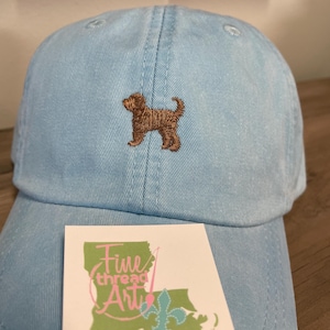 May include: A light blue baseball cap with a brown embroidered dog on the front. The cap has a white label with the text "Fine thread Art" and a green outline of the state of Louisiana with a pink needle and a fleur-de-lis.